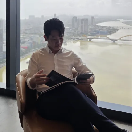 Ngô Đức Tính ngồi đọc sách bên cửa kính, phía sau là sông Hàn